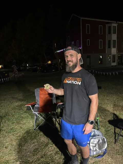 Bearded man in a backward cap, Mudder Nation T-shirt and blue shorts holds up a medal at night beside a camp chair