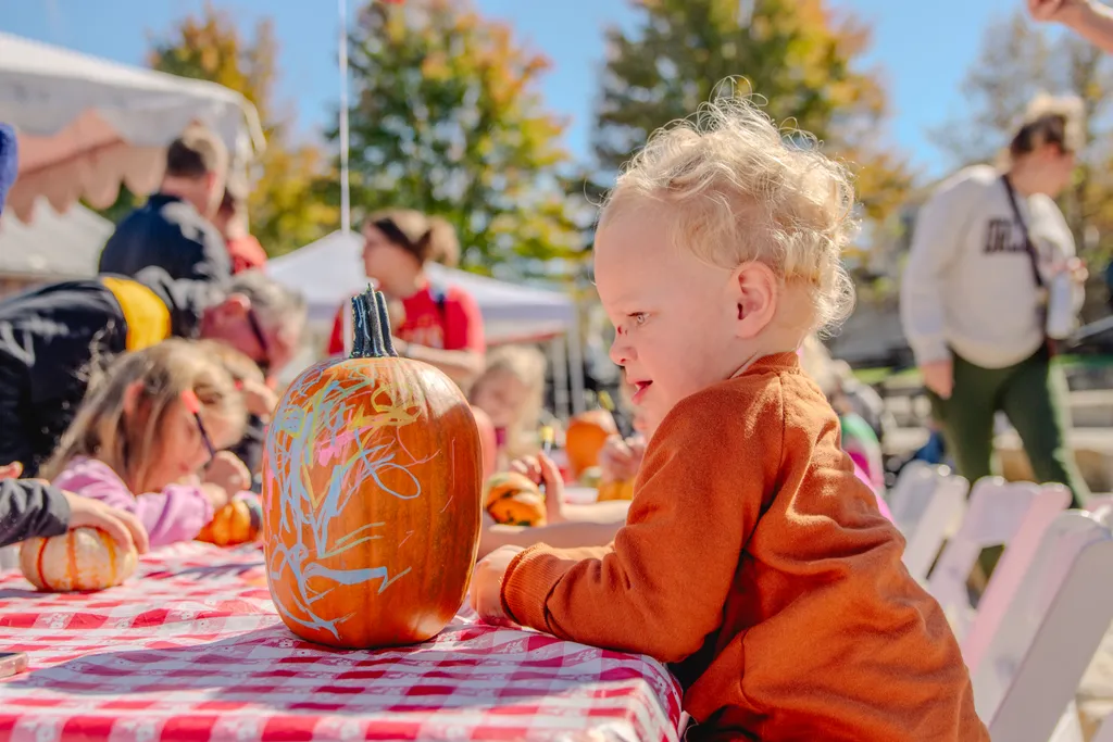 Curly-haired toddler in an orange sweater leans on a red checkered table beside a pumpkin scribbled with colorful chalk.