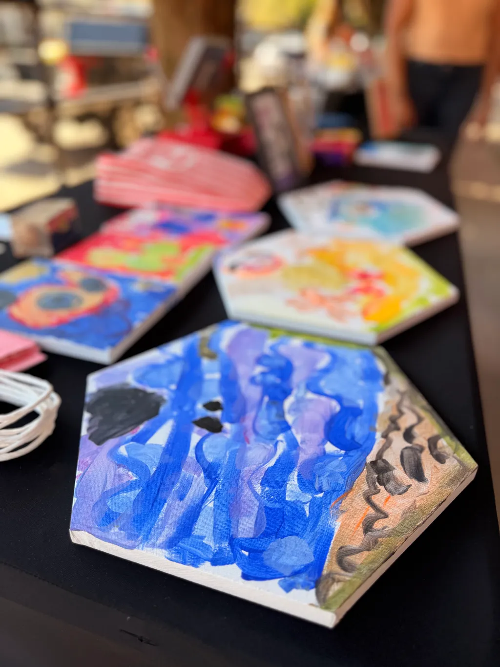 Hand-painted hexagon canvases in blue, purple, yellow and pink laid out on a black table, with blurred shoppers behind.