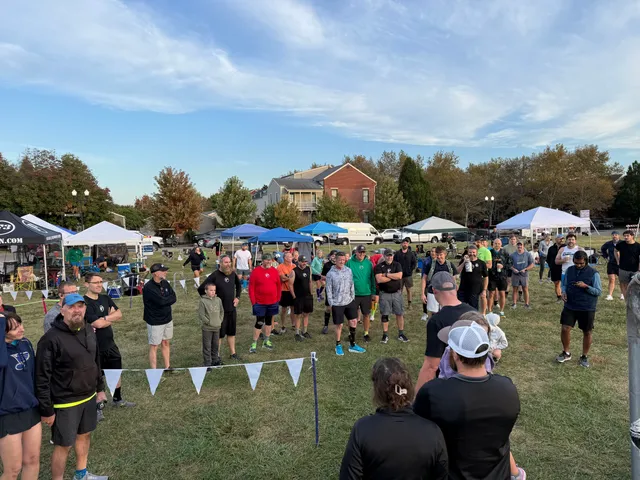 Runners in athletic gear gather in a circle on a grassy field with pop-up tents and white pennant flags under a blue sky