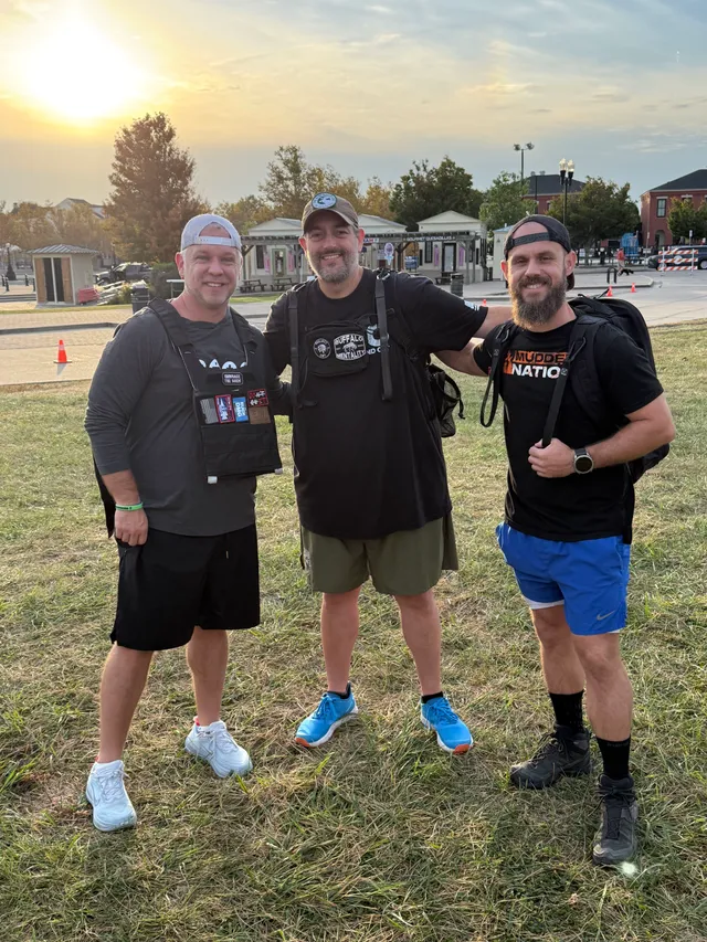 Three smiling men in black shirts, rucksacks and a weighted vest pose on grass at sunrise near park buildings.