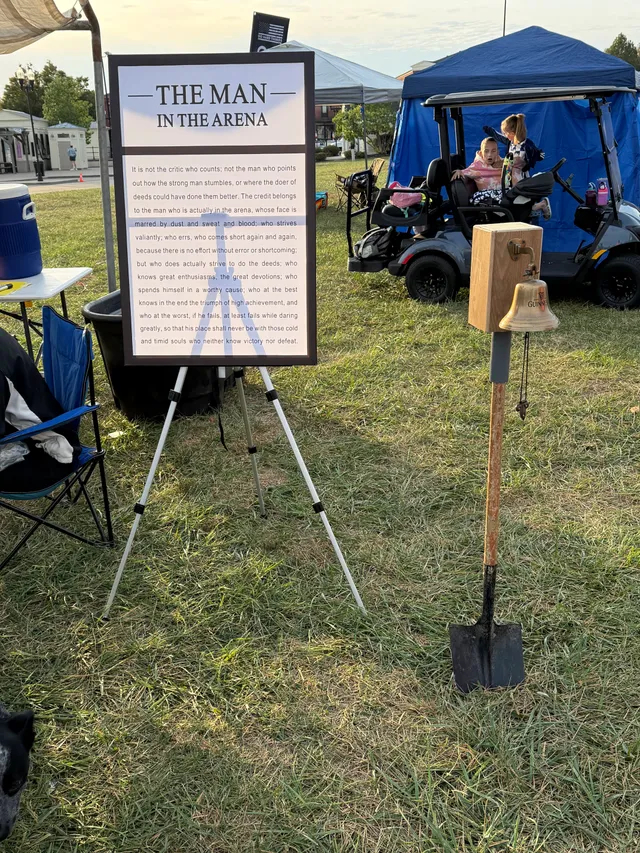 "The Man in the Arena" sign on an easel beside a brass bell mounted on a shovel, with kids in a golf cart near blue tents