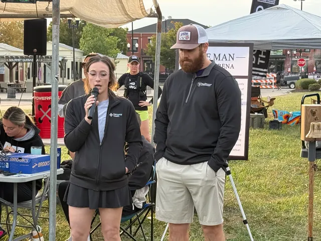 Woman in glasses speaks into a microphone beside a bearded man in a cap under a tent at an outdoor race event