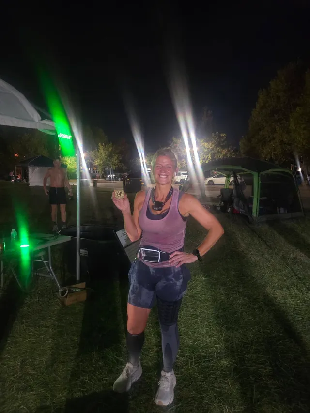 Smiling runner in a purple tank top and compression gear holds up a finisher coin on a grassy field at night near tents.