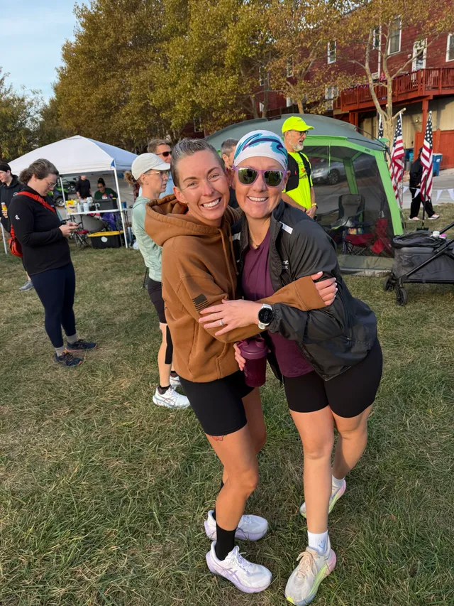 Two smiling women in running shorts and sneakers hug on grass near a green tent, canopy and American flags.