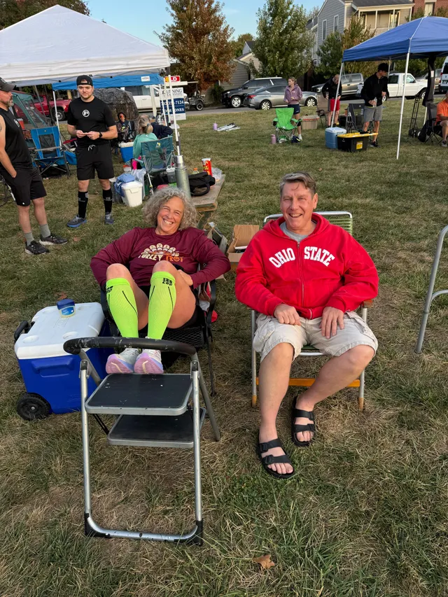 Smiling woman in neon green compression socks rests feet on step stool beside smiling man in red Ohio State hoodie on lawn