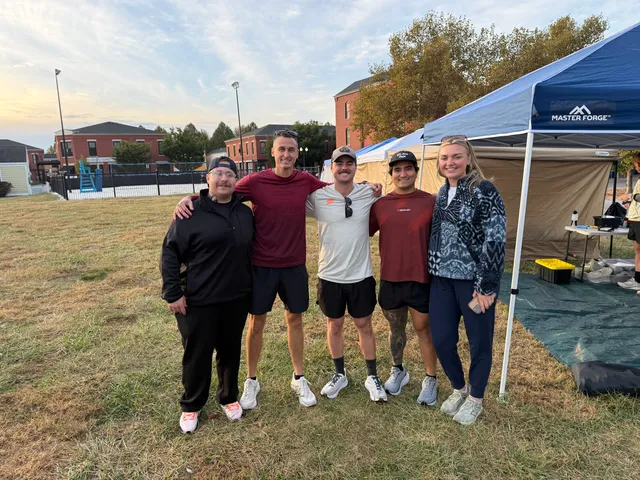 Five people in athletic wear pose arm in arm on grass beside a blue Master Forge canopy tent, brick buildings behind.