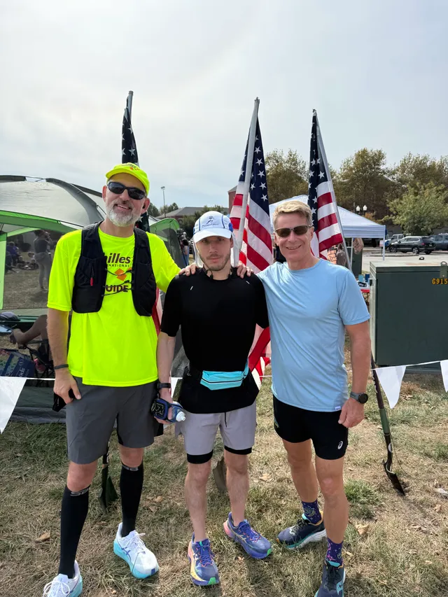 Three runners pose on grass in front of three American flags and a green canopy tent at an outdoor race camp.