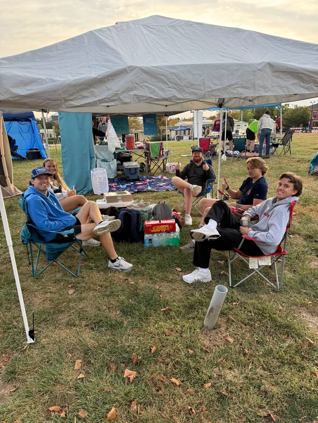 Five people in camp chairs under a gray pop-up canopy on grass, two giving thumbs up, with coolers and water cases