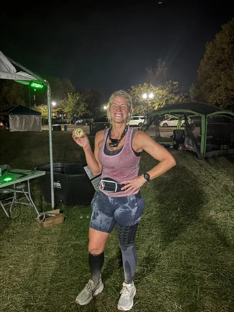 Smiling runner in pink tank, tie-dye shorts and compression socks holds up a small medal at a lit campsite at night