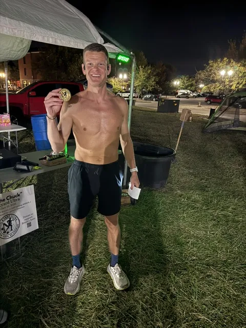 Shirtless runner in black shorts holds up a gold medal at night beside a canopy tent on grass, with a race sign nearby.
