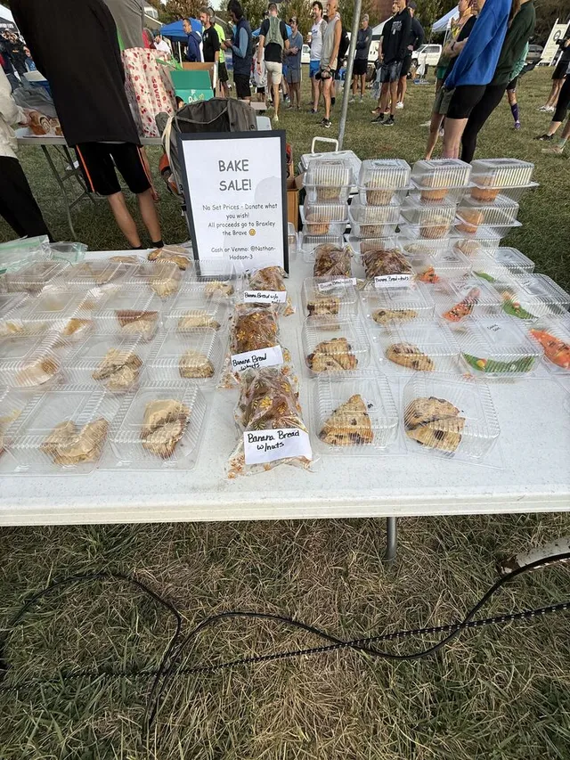 Outdoor table of baked goods in clear containers, banana bread bags, and a Bake Sale sign, with runners on grass behind