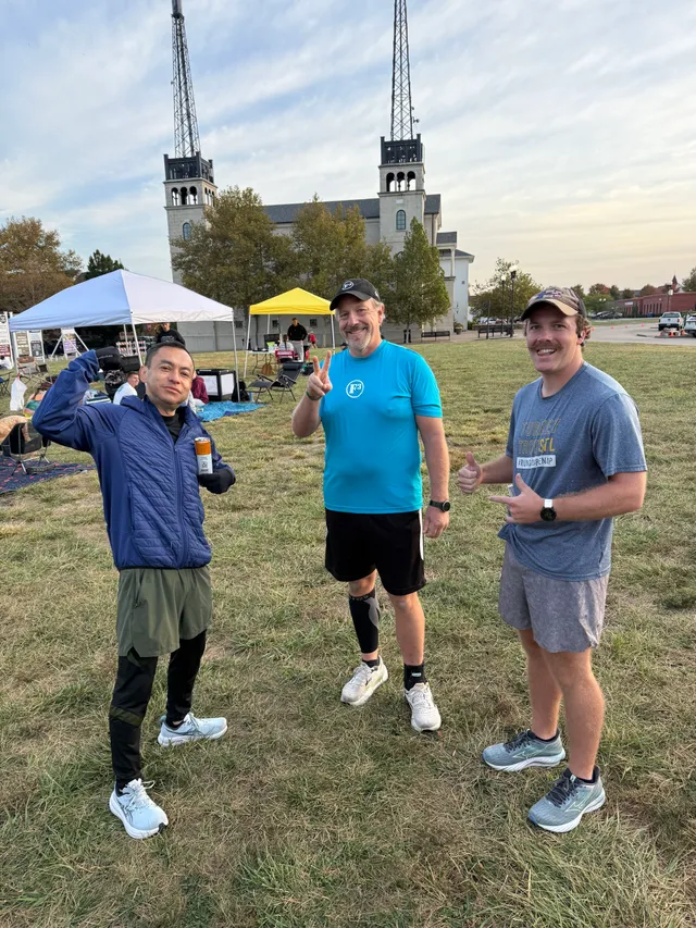 Three runners pose on a grassy field giving thumbs-up and peace signs, with canopy tents and a twin-towered building behind