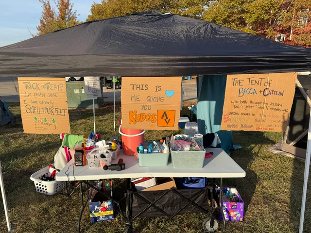 Black pop-up canopy over a folding table of runner supplies with handmade cardboard signs, one reading The Tent of Becca + Caitlin