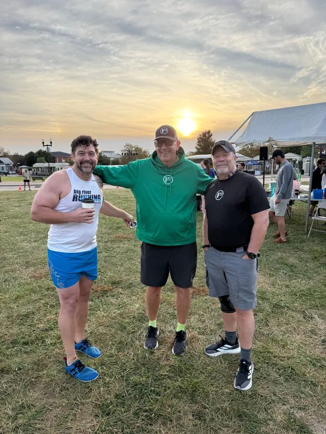 Three smiling men pose on grass at sunrise, one in a Big River Running tank holding coffee, near a white event tent.