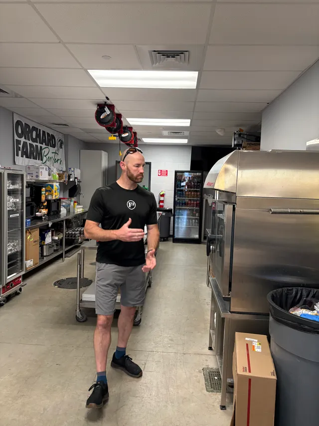 Man in black F3 shirt and gray shorts gestures inside a concession stand kitchen with steel ovens and an Orchard Farm Boosters banner