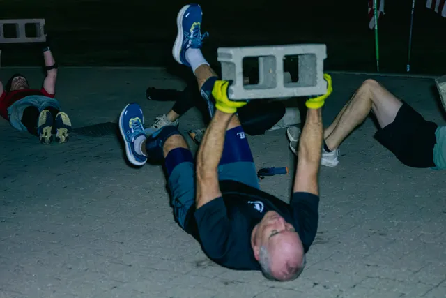 Man lying on pavement lifting a cinder block overhead with yellow gloves, legs raised, as others exercise nearby in the dark