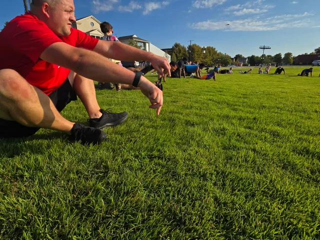 Man in red shirt sits on grass pointing down while adults and kids do plank exercises across a sunny field