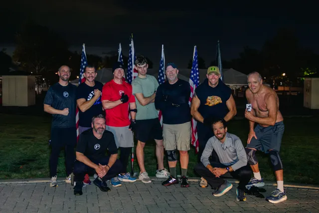 Nine men in workout clothes pose before dawn on a brick path, several flexing, with American flags behind them.