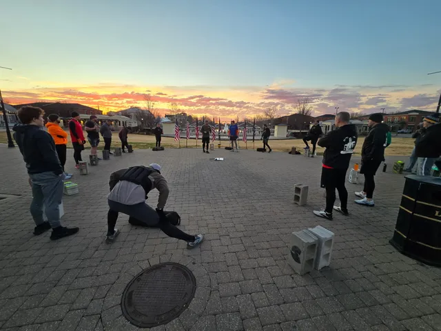 Men stand in a wide circle on a brick plaza at sunrise with cinder blocks, one stretching in a weighted vest, flags behind