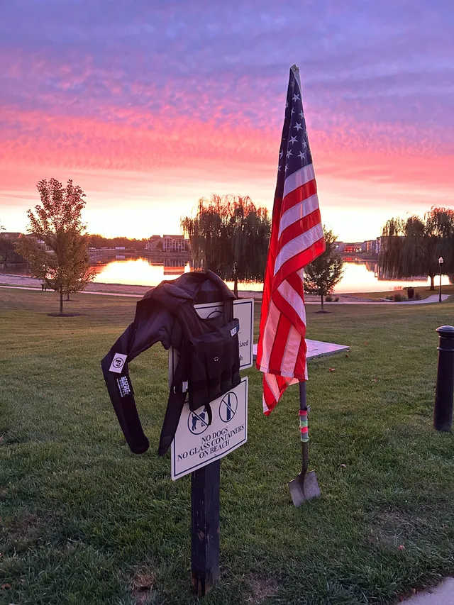 Black weighted vest hung on a 'No Dogs, No Glass Containers on Beach' sign beside an American flag on a shovel, pink sunrise over a lake