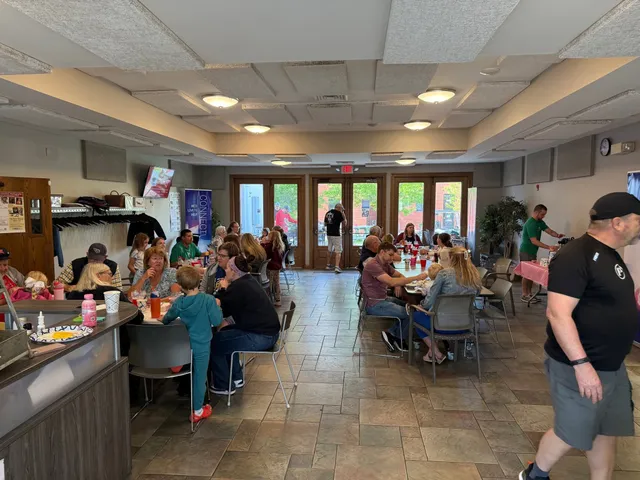 Families eat breakfast at long tables in a tiled community hall while men in black and green shirts walk and serve food.