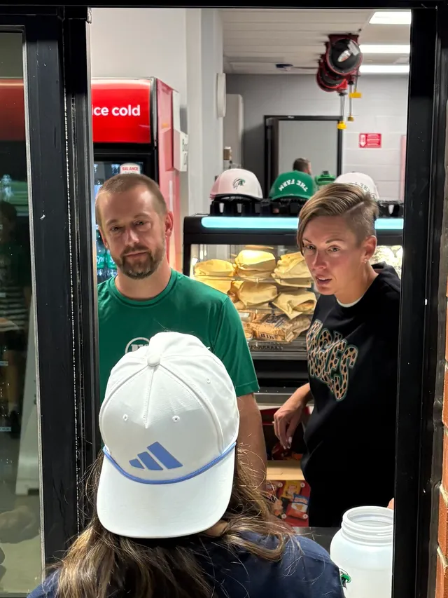Two volunteers in green and black shirts serve a customer in a white cap at a concession stand window with a sandwich warmer.