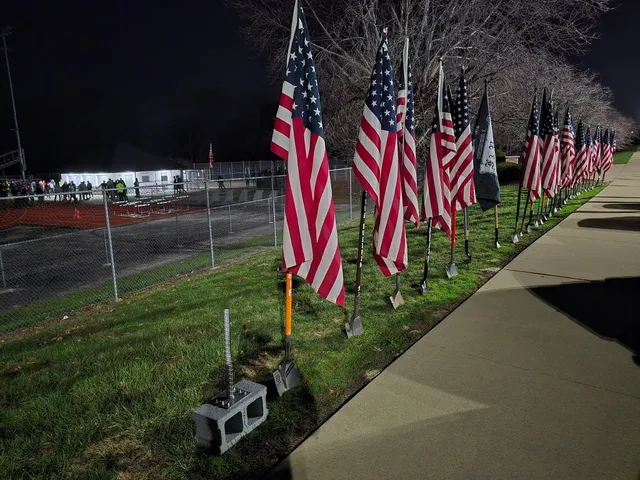 Row of American flags on shovel-handle poles along a sidewalk at night, beside a fenced track where people gather