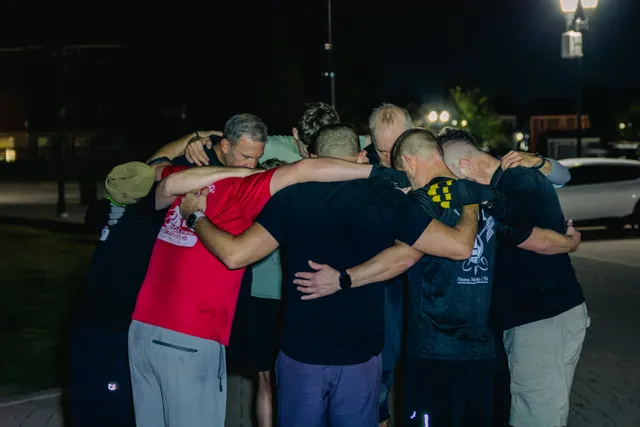 A group of men in workout clothes huddle with arms around each other's shoulders, heads bowed, outdoors at night.