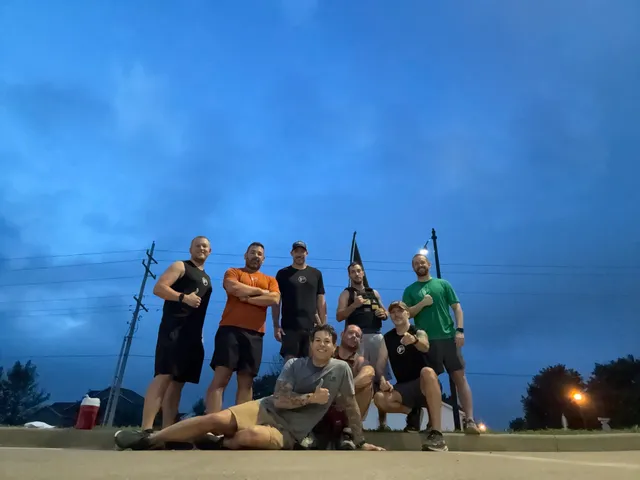 Eight men in workout clothes pose with thumbs up on a curb under a deep blue dawn sky, streetlights glowing