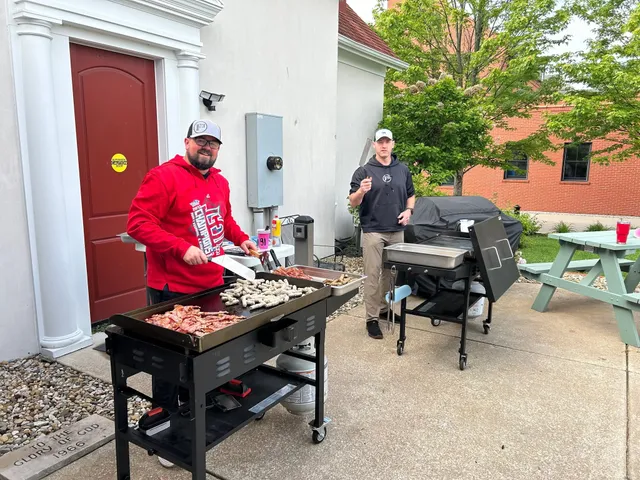 Man in red hoodie cooks bacon and sausages on a flat-top griddle outside a building as another man stands by a grill