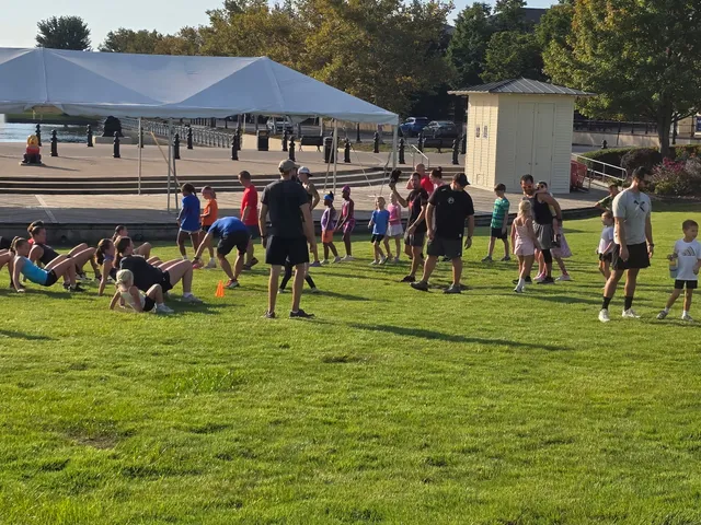 Adults and kids exercise on a grassy lawn; a row does sit-ups while others walk past a white tent and small shed by a lake.
