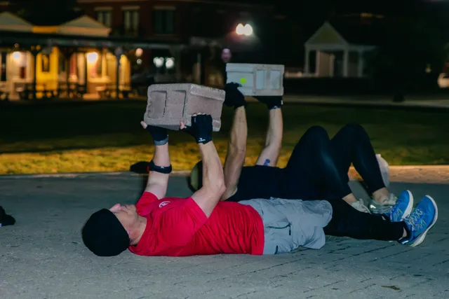 Two men lie on pavement at night pressing cinder blocks overhead, one in a red shirt and black beanie, lit buildings behind.