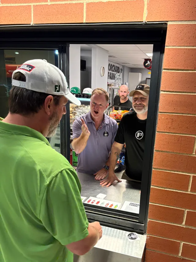 Man in green polo and white cap at a brick concession window; men in purple polo and black F3 shirts lean on the counter inside