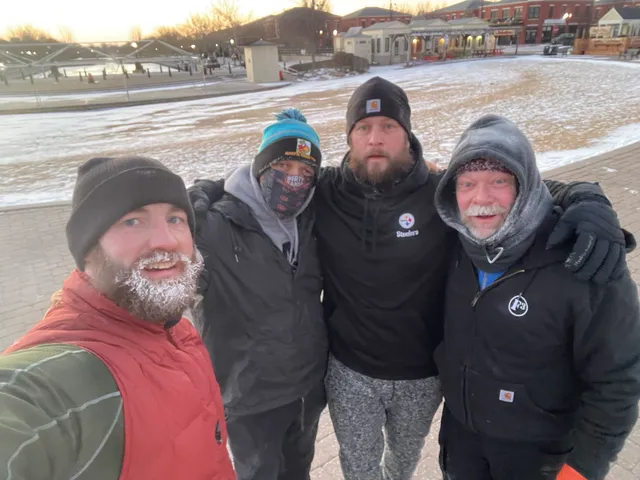 Four men in winter hats and dark jackets pose for a selfie on a snowy plaza at dawn, one with frost in his beard.