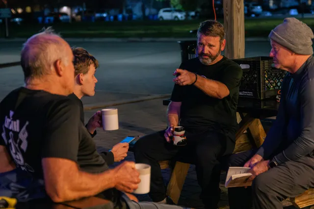 Four men sit at wooden picnic benches outdoors before dawn, holding coffee mugs as one bearded man gestures while talking.