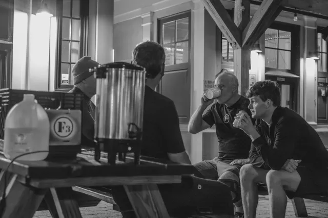 Black-and-white photo of four men at a picnic table drinking from cups beside a coffee urn and F3 crate outdoors.