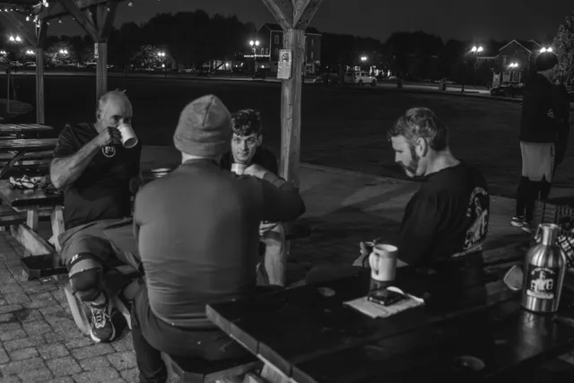 Black-and-white photo of men drinking coffee from mugs at picnic tables under a pavilion before dawn, streetlights behind.