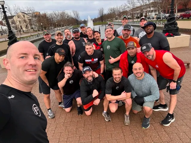 Selfie of about 20 men in workout clothes posing on a brick plaza by a pond with a fountain and lampposts