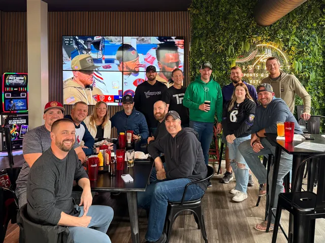 Group of about 14 people posing around bar tables at a sports bar, with football on a large video wall behind them.