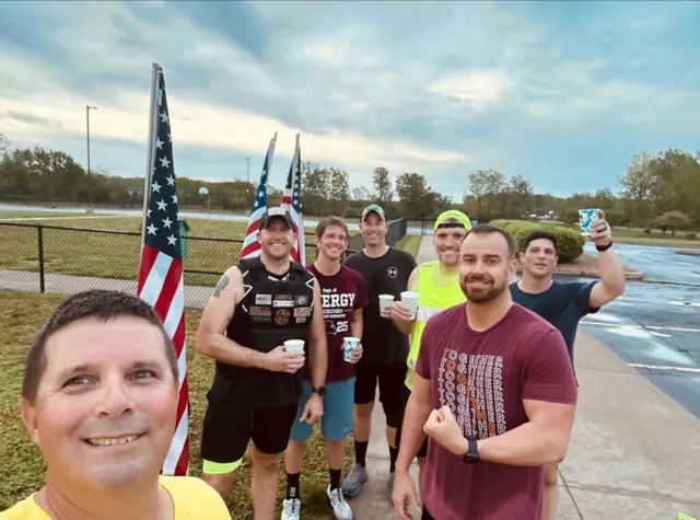 Seven men pose for a selfie on a sidewalk beside American flags, several raising paper cups, under a cloudy sky.