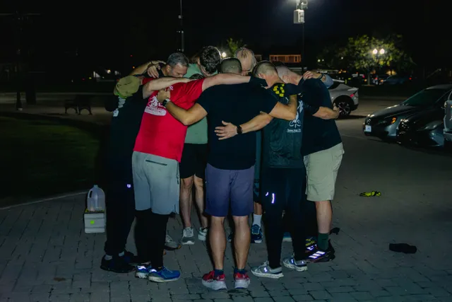Group of men in athletic shirts and shorts huddle with arms around each other on a brick plaza at night near parked cars