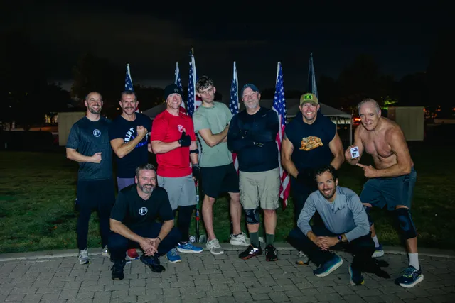 Nine men in workout clothes pose at night on a brick path in front of a row of American flags, some flexing.
