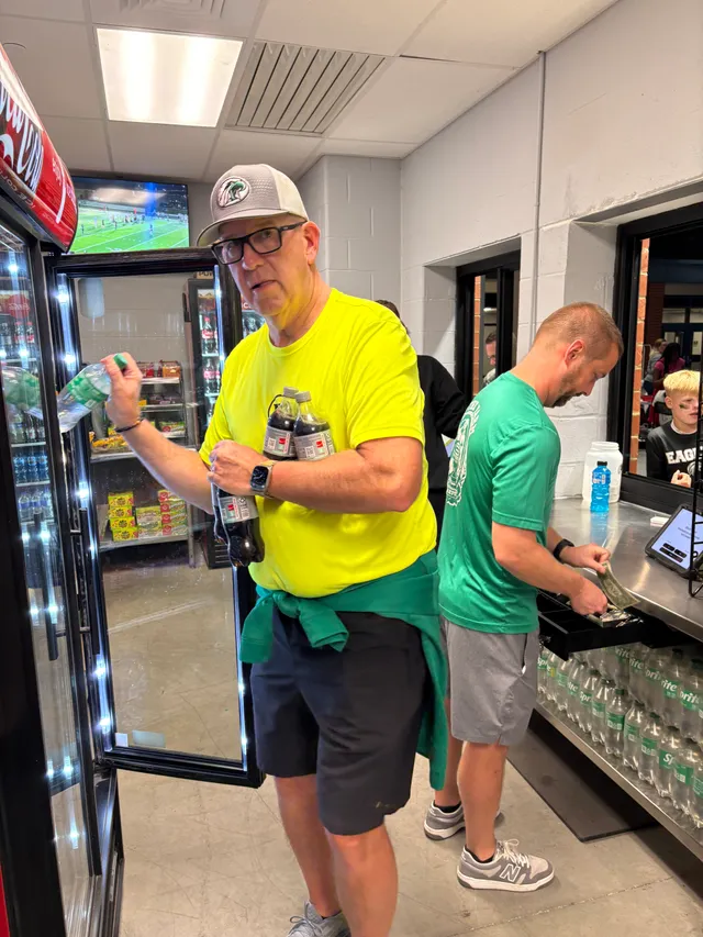 Man in neon yellow shirt and cap grabs bottles from a drink cooler while a man in green counts cash at a concession counter.