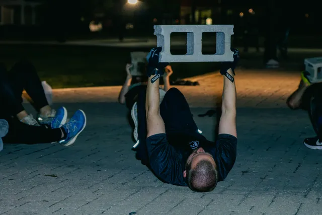 Man lying on brick pavement in the dark pressing a cinder block overhead, with others doing the same nearby