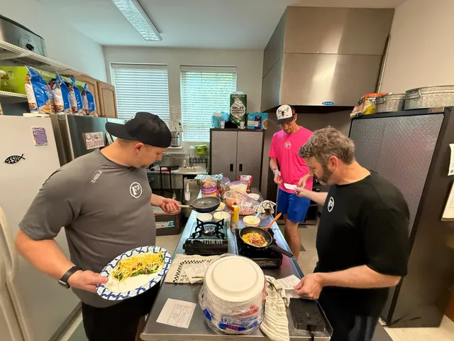 Three men cook on portable burners at a stainless steel table in a kitchen, one holding a plate of greens and cheese.