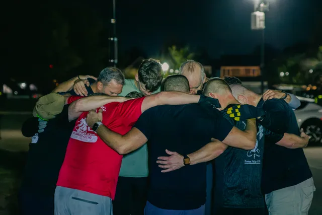 A group of men in workout clothes huddle with arms around each other, heads bowed, in a dark parking lot under lights.