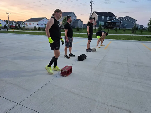 Four men in black workout shirts stand by cinder blocks in an empty parking lot at sunrise, houses behind them