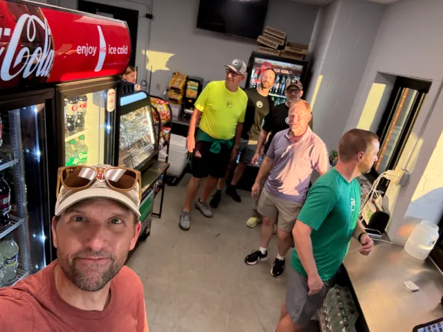 Man in a cap and sunglasses takes a selfie with five men in a concession stand with Coca-Cola coolers and snack displays