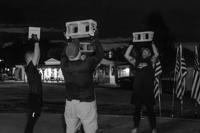 Black-and-white photo of men lifting cinder blocks and a paver overhead at night beside American flags